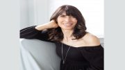 A woman with long dark hair, wearing a black off-shoulder top and a necklace, is smiling and posing with her hand resting on the back of her neck, excitedly discussing how the Cookbook Club returns at Milton Public Library on June 5.