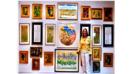 A woman in a yellow shirt and white pants stands beside a gallery wall filled with various framed floral artworks, while eagerly discussing the upcoming "Wildlife of the Serengeti" virtual safari talk with Joy Marzolf on June 18, 2024.