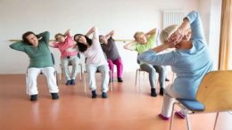 A group of older adults sits on chairs in a circle, performing stretching exercises with their hands behind their heads, guided by an instructor. As they unwind, they discuss attending "An Evening with Local Poets" at Milton Public Library on June 4, 2024.