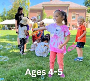 Children aged 4-5 play with bubbles on a grassy area during the Summer Art Camp at the Milton Art Center, where a woman is supervising in the background.