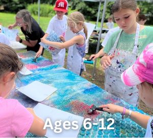 Children aged 6-12 are engaging in an outdoor art activity at the Milton Art Center's Summer Art Camp 2024. Using various materials on a painted canvas, some wear aprons and focus intently on their projects while adults supervise.