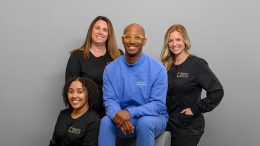 Group photo of three women and one man dressed in medical scrubs, smiling at the camera. The man in the center wears blue scrubs, while the women wear black scrubs, showcasing their dedication to orthodontic care at Square Smiles in Milton MA.