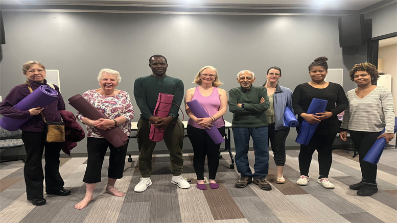 A group of seven people stands in a room at the Milton Public Library, each holding a yoga mat and smiling at the camera. The floor is carpeted, and the walls are plain with minimal decor, as part of their adult programming for July 2024.