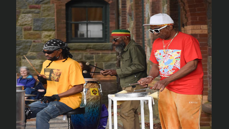 Three musicians wearing colorful clothing are performing outdoors. One plays a drum set, another plays congas, and the third plays a steelpan. A brick and stone building stands in the background. Experience holiday magic at Eustis Estate on December 14: an evening of festive cheer awaits you there!