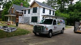 A two-story house under renovation in 2025, with Tyvek sheathing on the exterior, stands ready for transformation. A white van with the Costa Painting Services logo and a ladder on top is parked in the driveway. Construction materials are scattered nearby, hinting at the magic of a fresh coat of paint to come.