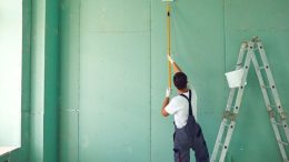 A person in overalls and gloves paints a green wall using a paint roller on an extended handle. A ladder and a small bucket are nearby, representing the expertise of Costa Painting Services Inc: Excellence in painting since 2007.