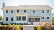 Discover excellence in architecture with MASS Architect. This large, two-story house features light blue siding, multiple windows, a chimney, and a back porch that includes a barbecue grill and outdoor furniture, all surrounded by a meticulously landscaped yard.