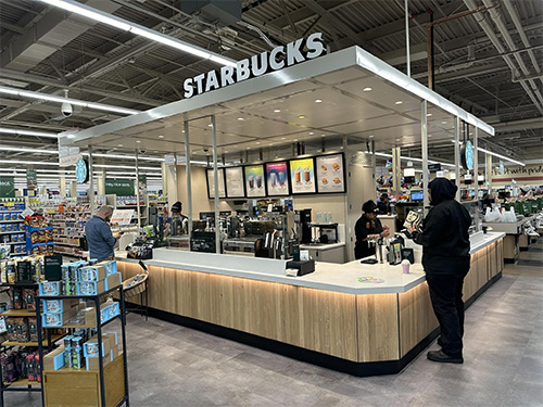 A Starbucks counter located within a grocery store, where several customers are being served by baristas. Shelves with various products surround the area, creating a busy but inviting atmosphere. Nearby, a sign reminds everyone of Cunningham Woods dog rules to ensure a pleasant experience for all.