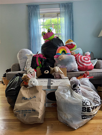 A living room with a sofa, piled high with various stuffed animals and bags of donations, including labeled shopping bags and black trash bags. A window with blue curtains is in the background, suggesting a community spirit reminiscent of Top Milton Neighbors posts.