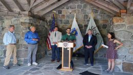 A man speaks at a podium in an outdoor stone pavilion, surrounded by five people, during the St. Patrick’s Family Celebration and Fundraiser event to take place March 11. Flags and a stone wall are in the background.