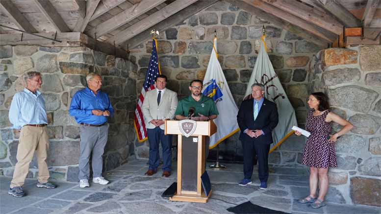 A man speaks at a podium in an outdoor stone pavilion, surrounded by five people, during the St. Patrick’s Family Celebration and Fundraiser event to take place March 11. Flags and a stone wall are in the background.