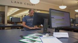 A person in a "Foxboro" t-shirt uses a computer in the Land Court room. In the foreground, a desk displays documents labeled "Ancestry Research." Meanwhile, Register O’Donnell announces re-election bid, highlighting achievements and future plans.