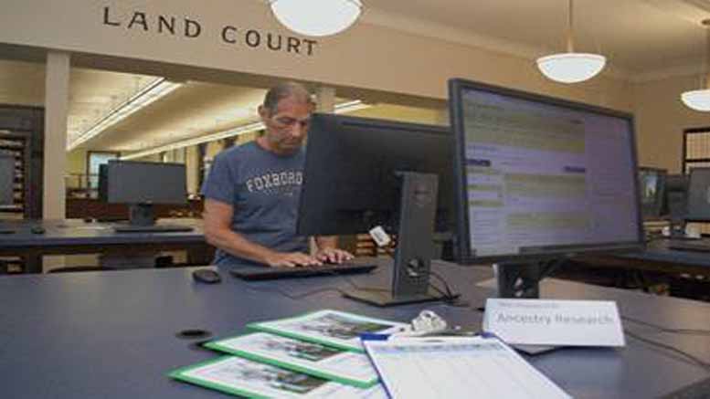 A person in a "Foxboro" t-shirt uses a computer in the Land Court room. In the foreground, a desk displays documents labeled "Ancestry Research." Meanwhile, Register O’Donnell announces re-election bid, highlighting achievements and future plans.