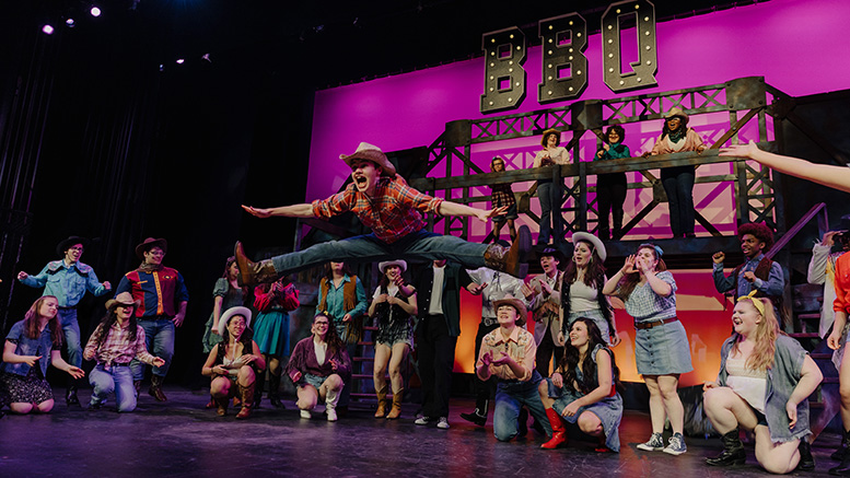 A group of performers on stage, many dressed in western attire, with one person in mid-air doing a split jump. The background features a large sign that reads "BBQ." This lively act is brought to you by the Milton Neighbors Choice Award-winning Boxcar Stage Company.