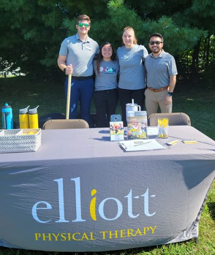 Four people stand behind a table adorned with an "Elliott Physical Therapy" tablecloth, brochures, pens, and water bottles. The setup is outdoors next to a tree. Proudly displayed is the Milton Neighbors Choice Award 2024 banner that showcases their community recognition.