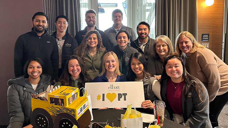 A diverse group of 13 people smiling, gathered around a table. They are holding a sign that reads "#I ❤️ PT" with a model of a yellow truck on the table. This joyful moment was captured at Elliott Physical Therapy, proud Milton Neighbors Choice Award Winner.