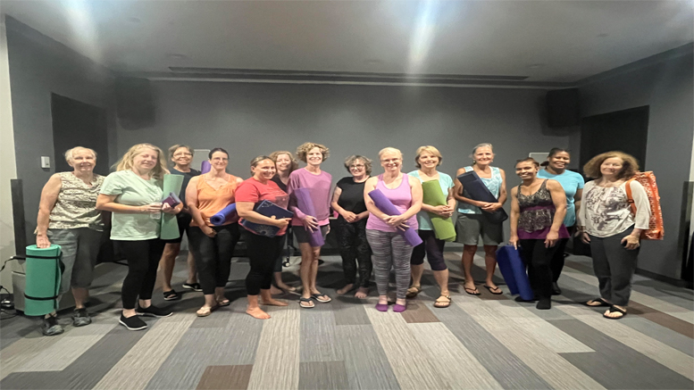 A group of fifteen people stands indoors, holding rolled-up yoga mats and smiling at the camera. They appear to be in a yoga studio or similar setting, likely gearing up for the Free Adult Yoga class at Milton Public Library on August 6, 2024.