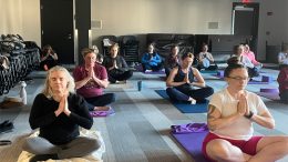 A group of people are seated on yoga mats in a calm room, with eyes closed and hands in a prayer position, participating in a yoga meditation session. Milton Public Library announces adult programs for September 2024, including sessions like this to enhance well-being.