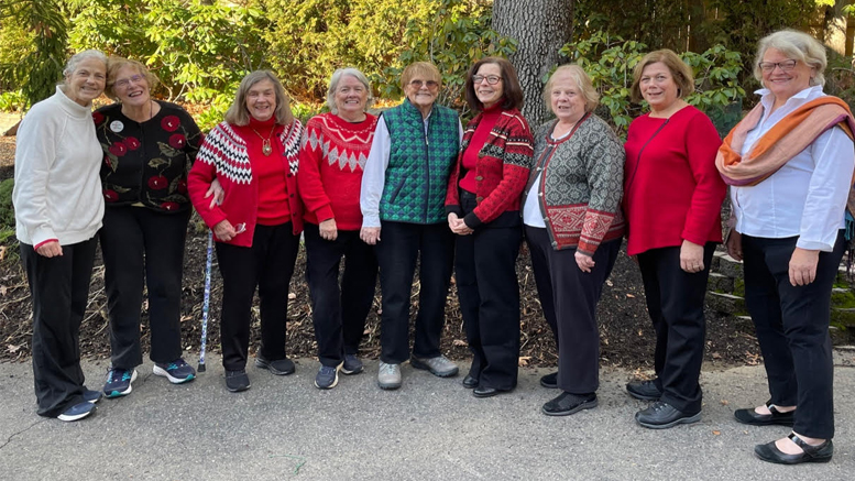 Nine elderly women, some wearing red or patterned sweaters, stand together outdoors, smiling at the camera on a paved path with trees in the background. Nearby, the Milton Public Library showcases Andrise Dalmace’s vibrant floral paintings in Wotiz Gallery throughout August.