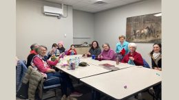 A group of people sits around a table in a room at the Milton Public Library, engaging in various activities. The background features a painting on the wall, and an announcement highlights adult programs for September 2024.