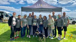 A group of people from Elliott Physical thearpy wearing matching gray shirts stands together in front of an Elliott Physical Therapy tent, showing off their numbered bibs at a race event. Proudly displayed is a banner announcing Elliott Physical Therapy as the 2024 Milton Neighbors Choice Award Winner.