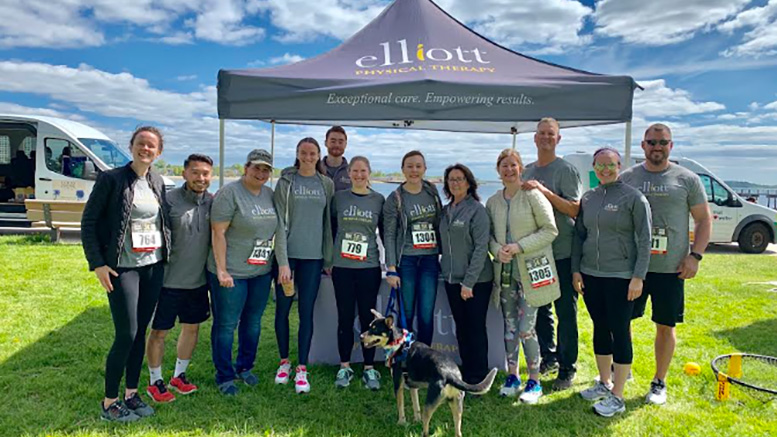 A group of people from Elliott Physical thearpy wearing matching gray shirts stands together in front of an Elliott Physical Therapy tent, showing off their numbered bibs at a race event. Proudly displayed is a banner announcing Elliott Physical Therapy as the 2024 Milton Neighbors Choice Award Winner.