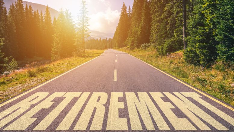 A road extends into a forest with the word "RETIREMENT" written in large letters on the pavement, illuminated by sunlight breaking through the trees, inviting those who retire to embark happily on a serene journey.