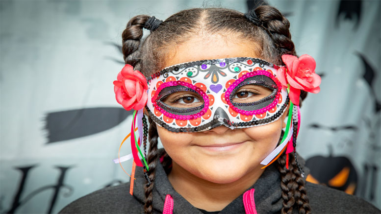 A girl with braided hair, adorned with a colorful mask featuring floral decorations and two pink flowers in her hair, participates in Interfaith Social Services collecting Halloween costumes for 2022.