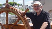 A man wearing glasses and a cap stands next to a large ship's steering wheel labeled "Cutty Sark." He is smiling, with one hand on the wheel, as Milton Public Library announces adult programs for October 2024.