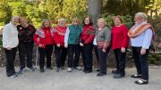 A group of nine elderly women standing outdoors, wearing sweaters and jackets, smiling at the camera. Trees and bushes are visible in the background, as they enjoy the Winter Light. This joyful moment is part of the upcoming Wotiz Gallery exhibit to take place in March.