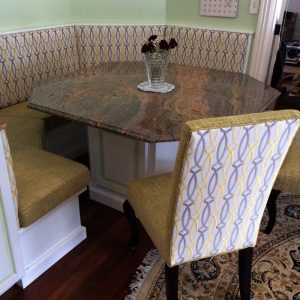 Upholstery by Michael: A kitchen nook with green cushioned seating and a large granite table, featuring elegant upholstery. A vase with dark red flowers sits at the center of the table, while the surrounding chairs boast a striking geometric pattern.