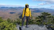 A person in a yellow jacket, hiking pants, and boots stands on a rocky outcrop with a scenic view of mountains and a forested valley in the background, reminiscent of the vistas celebrated by Friends of the Blue Hills during their Annual Celebration on October 4th.