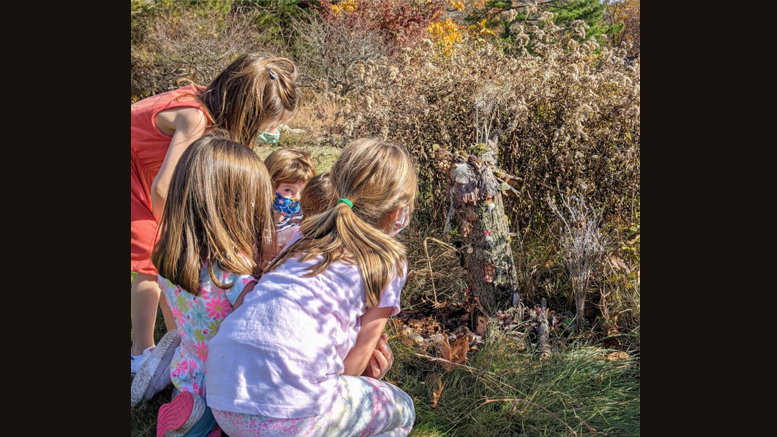 Four children crouch in a grassy area, imagining fairies in the foliage as they gaze intently at a small makeshift structure on the ground, surrounded by dried plants. Consider exploring similar wonders at the Eustis Estate during their self-guided tours from Nov. 5-12.