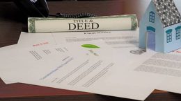 Documents, including a title and deed, are spread on a table next to a small paper house model, under the watchful eye of Register O’Donnell at the Norfolk County Registry's newly launched free genealogy research center.