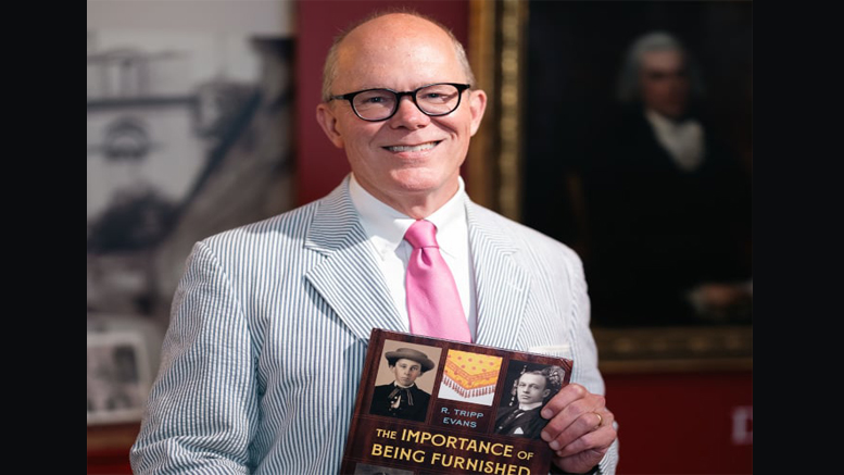 A man in a striped suit and pink tie holds a book titled "The Importance of Being Furnished" while standing in front of framed pictures, reminiscent of the elegance found at the Eustis Estate's newest exhibition, "Changing Landscape: Sculpture.