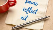 A red mug and a silver pen rest on a napkin with the words "pause, reflect, act" written in blue—a mantra for setting new goals as you craft a winning marketing strategy for 2024.