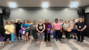 A group of people stands in a room at the Milton Public Library, each holding a yoga mat and posing for a photo, celebrating the announcement of adult programs set for December 2024.