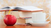 An open book rests atop two closed ones beside a red apple and a white cup with saucer on the wooden table, setting the perfect scene for Booked for Lunch: Reading & Socializing on November 16, 2024.