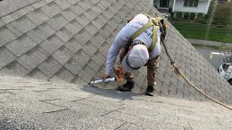 Wearing safety gear, a person applies sealant to roof shingles on a sloped rooftop, taking essential steps to winter-proof your home and avoid costly repairs.