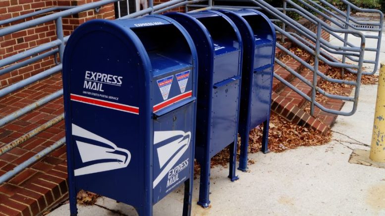 Three blue USPS mailboxes labeled "Express Mail" are lined up on a sidewalk near a brick building and silver railings, while the Milton Police Department investigates reports of stolen checks.