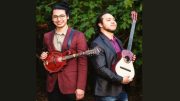 Amid leafy greenery, Ethan Setiawan and Maurizio Fiore Salas stand back to back at their Eustis Estate concert. One holds a mandolin, the other a small guitar, creating an elegant tableau of strings and harmony.