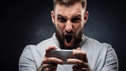 Man with a beard, wearing a light gray shirt, intensely focused and shouting while holding a gaming controller against a dark background. Perhaps it's the dopamine similar to why you're participating in heated conversations on social media.