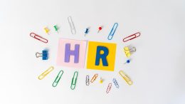 Colorful paperclips, push pins, and binder clips surround "HR" in bold letters on pink and yellow backgrounds, highlighting HR best practices essential for effective management in large workplaces.