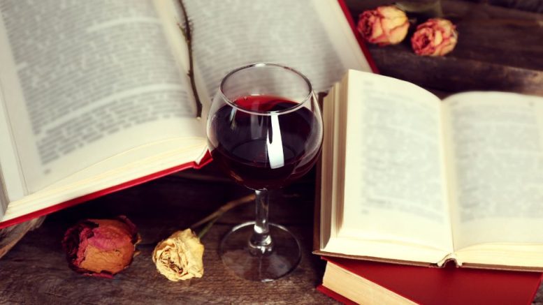 A glass of red wine sits on a wooden table amidst open books and dried roses, setting the perfect scene for Boozy Bookworms: Join the Under-40 Book Group – January 23, 2025.