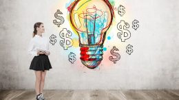 A woman stands holding a tablet, gazing at a wall adorned with a colorful lightbulb drawing surrounded by dollar signs. She's exploring "Small Business Corner's" top 3 cost-effective marketing ideas to kickstart your business in the new year.