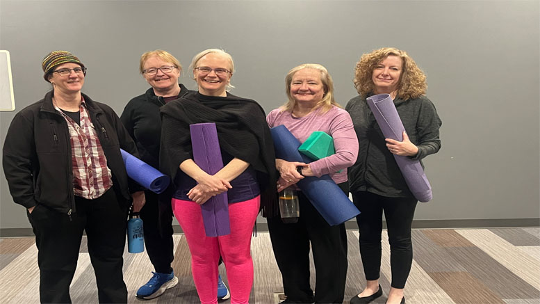 Five people stand indoors at the Milton Public Library, holding yoga mats and smiling at the camera, as they kick off the adult programs announced for March 2025.