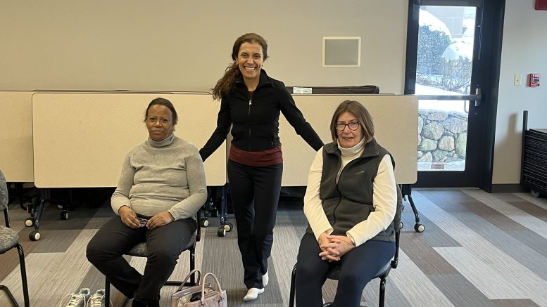 Three women in a room with two seated on chairs and one standing, all smiling. The setting includes beige folding tables and a patterned carpet, with shoes and a bag scattered on the floor—a glimpse into moments at the Milton Public Library's adult programs for March 2025.