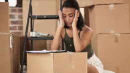 A woman sits on the floor with her hands on her temples, overwhelmed by relocation challenges, surrounded by moving boxes and papers strewn over the box in front of her. A ladder stands in the background, a silent reminder of how a move can go wrong at any moment.