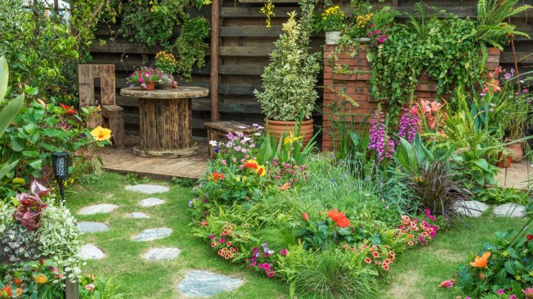 stunning gardens in a lush scene featuring a stone path, vibrant flowers, and greenery. A wooden table and chairs sit cozily in the corner, while a wooden fence entwined with climbing plants