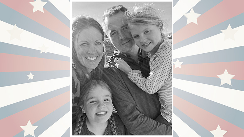 Smiling family of four, including two children, posing in front of a backdrop with red, white, and blue stars and stripes.